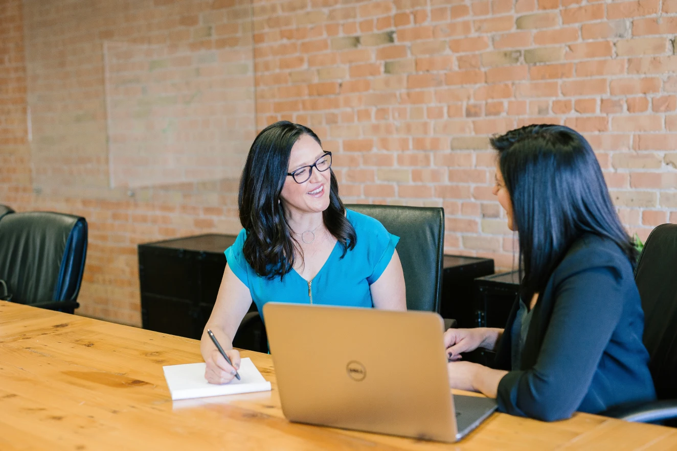 Two women discussing work, with one using a laptop and the other holding a pen.
