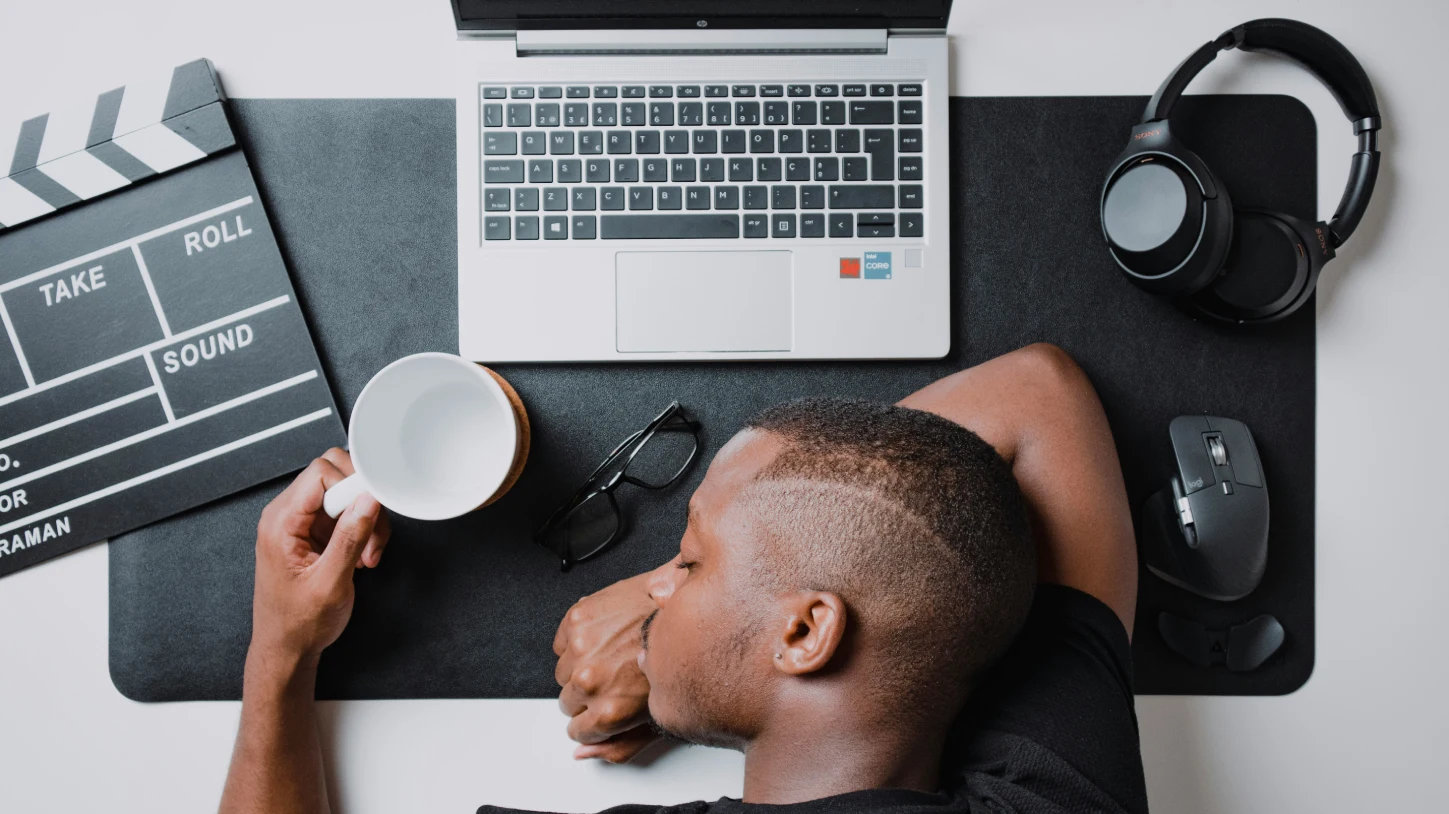 Man asleep at a desk while holding a coffee mug beside a laptop.